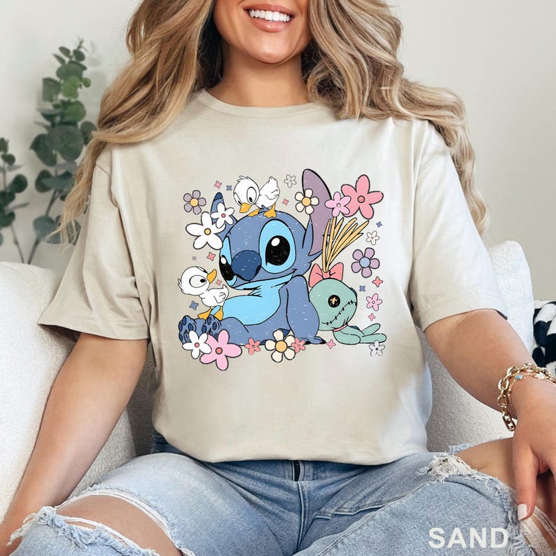 K&ouml;nnte beinhalten: Sandfarbenes T-Shirt mit einer blauen Cartoonfigur mit gro&szlig;en Augen, umgeben von Blumen und kleinen wei&szlig;en V&ouml;geln. Die Grafik enth&auml;lt eine gr&uuml;ne Puppe mit einer rosa Schleife. Das Wort "SAND" ist unten aufgedruckt.
