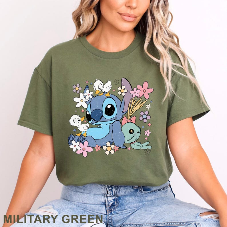 K&ouml;nnte beinhalten: Milit&auml;rgr&uuml;nes T-Shirt mit einer Cartoonfigur mit blauem Fell, gro&szlig;en schwarzen Augen und wei&szlig;en Entenk&uuml;ken. Das Design umfasst eine gr&uuml;ne Puppe und bunte Blumen. Der Text "MILITARY GREEN" befindet sich unten.