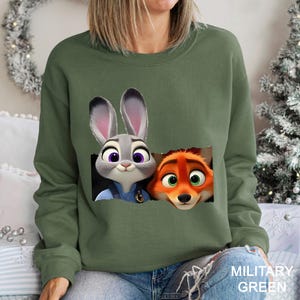 Może przedstawiać: Zielona wojskowa bluza z nadrukiem Judy Hopps i Nicka Wilde z Zootopii. Bluza ma okrągły dekolt i długie rękawy. Tekst "MILITARY GREEN" jest wydrukowany w prawym dolnym rogu obrazu.