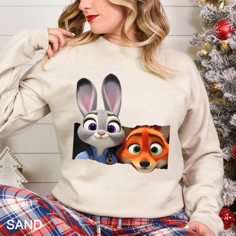 Może przedstawiać: Beżowa bluza z nadrukiem Judy Hopps i Nicka Wilde z filmu Zwierzogr&oacute;d. Postacie z kresk&oacute;wek są przedstawione jako wyglądające z podartego prostokąta. Bluza ma długie rękawy i okrągły dekolt. Słowo "SAND" jest wydrukowane na dole.