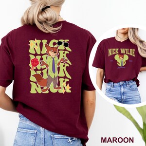 May include: Maroon t-shirt featuring a cartoon fox character with sunglasses and the name "Nick" in a stylized font. The character is wearing a green shirt and purple tie, holding a lollipop. The shirt also has the text "Nick Wilde" and "Zootopia".