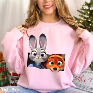 Może przedstawiać: Jasnor&oacute;żowa bluza z nadrukiem Judy Hopps i Nicka Wilde z filmu Zwierzogr&oacute;d. Postacie są przedstawione w prostokątnym wycięciu. Tekst "Light Pink" znajduje się w lewym dolnym rogu obrazu.