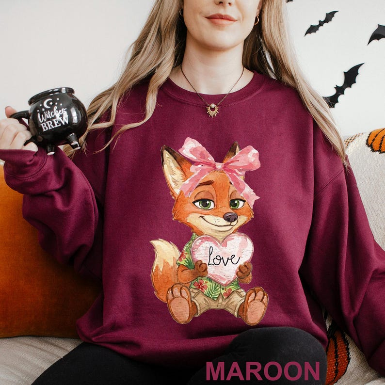 May include: Maroon sweatshirt with a cartoon fox holding a heart that says "Love." The fox has a pink bow and a Hawaiian shirt. The word "MAROON" is printed at the bottom of the sweatshirt.