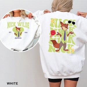 K&ouml;nnte beinhalten: Wei&szlig;es Sweatshirt mit einem Cartoon-Bild von Nick Wilde aus Zootopia. Auf der Vorderseite befindet sich ein kreisf&ouml;rmiges Design mit dem Text "Nick Wilde" und "Zootopia". Auf der R&uuml;ckseite ist ein gr&ouml;&szlig;eres Bild von Nick Wilde mit Sonnenbrille und Eis am Stiel zu sehen, umgeben von gr&uuml;nem Text.