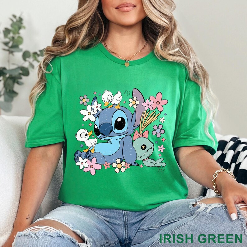 K&ouml;nnte beinhalten: Gr&uuml;nes T-Shirt mit einer Cartoonfigur mit blauer Fellfarbe, gro&szlig;en Augen und Entenk&uuml;ken. Das Design umfasst rosa, wei&szlig;e und lila Blumen. Der Text "IRISH GREEN" befindet sich am unteren Rand des T-Shirts.