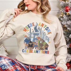 May include: Sand-colored sweatshirt featuring a colorful graphic of a city skyline and various animal characters. The text "WHERE ANYONE CAN BE ANYTHING" is displayed above the characters. The sweatshirt has long sleeves.