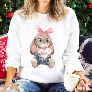 May include: White crew neck sweatshirt with a cartoon bunny holding a heart-shaped sign with the word "Love". The bunny has a pink bow and is wearing a police uniform. The sweatshirt is a casual style.