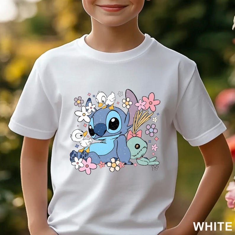 K&ouml;nnte beinhalten: Wei&szlig;es T-Shirt mit einer Cartoon-Illustration eines blauen, au&szlig;erirdisch aussehenden Charakters, umgeben von Blumen und kleinen Entenk&uuml;ken. Ein gr&uuml;ner puppenartiger Charakter mit einer rosa Schleife ist ebenfalls abgebildet. Das Wort "WHITE" befindet sich unten rechts.
