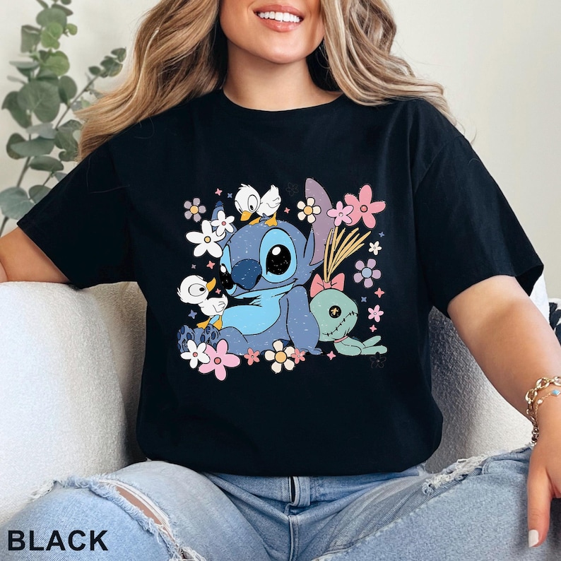 K&ouml;nnte beinhalten: Schwarzes T-Shirt mit einer Cartoon-Illustration von Stitch, umgeben von Blumen und zwei Entenk&uuml;ken. Eine gr&uuml;ne Puppe ist ebenfalls abgebildet. Das Wort "BLACK" ist unten auf dem T-Shirt aufgedruckt.