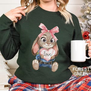 May include: A forest green sweatshirt with a cartoon bunny holding a heart-shaped sign with the word "Love." The bunny has a pink bow and is wearing a blue uniform. A white mug is held in the hand.