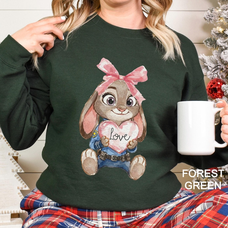 May include: A forest green sweatshirt with a cartoon bunny holding a heart-shaped sign with the word "Love." The bunny has a pink bow and is wearing a blue uniform. A white mug is held in the hand.