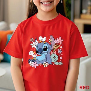 K&ouml;nnte beinhalten: Rotes T-Shirt mit einem Cartoon-Motiv von Stitch, umgeben von Blumen und Enten. Eine gr&uuml;ne Puppe mit rosa Schleife ist ebenfalls abgebildet. Das Wort "RED" ist unten rechts aufgedruckt. Kinder-T-Shirt.