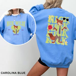 K&ouml;nnte beinhalten: Carolina-blaues Sweatshirt mit einem Cartoon-Fuchs-Charakter und dem Text "Nick Wilde". Auf der R&uuml;ckseite des Sweatshirts befindet sich ein Cartoon-Fuchs mit Sonnenbrille und dem Text "Nick Wilde". Die Vorderseite hat ein kreisf&ouml;rmiges Design.
