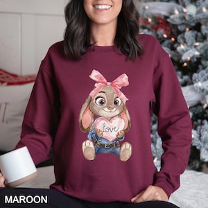 May include: Maroon sweatshirt with a cartoon bunny holding a heart-shaped sign with the word "Love". The bunny wears a pink bow and a police uniform. The word "MAROON" is printed at the bottom of the image.