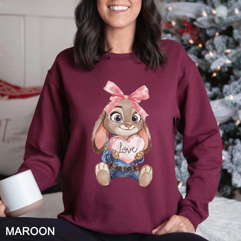 May include: Maroon sweatshirt with a cartoon bunny holding a heart-shaped sign with the word "Love". The bunny wears a pink bow and a police uniform. The word "MAROON" is printed at the bottom of the image.