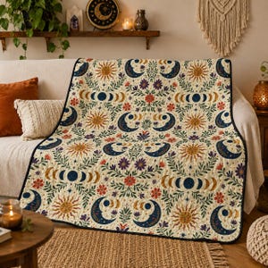 May include: A quilted throw blanket featuring a repeating pattern of suns, crescent moons, and lunar phases interspersed with colorful floral and leafy vine motifs on a cream background, finished with a dark blue stitched border.