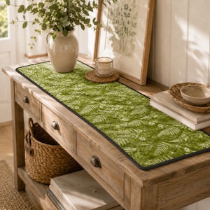 May include: A rectangular quilted table runner featuring a vibrant green background with a white botanical pattern of ferns and small flowers. The runner has a dark black piped edge and is displayed on a wooden console table with drawers.