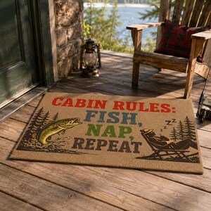 Könnte beinhalten: Eine rechteckige, hellbraune Fußmatte mit dem Text CABIN RULES: FISH, NAP, REPEAT in Rot, Blau und Grün. Das Design zeigt links einen springenden Fisch und rechts eine in einem Stuhl schlafende Person, umgeben von Tannenbäumen.