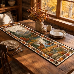 May include: A rectangular quilted table runner featuring a printed landscape scene of a deer in a forest with mountains, surrounded by a border of autumn leaves and pinecones in shades of orange, yellow, brown, and green on a wooden table.