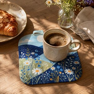 Könnte beinhalten: Ein quadratischer Topflappen aus Stoff mit einem Druck von sanften Hügeln, weißen Gänseblümchen und blauen Wildblumen. Eine gesprenkelte, beige Keramiktasse mit schwarzem Kaffee steht auf dem Topflappen, der oben eine kleine Stoffschlaufe besitzt.
