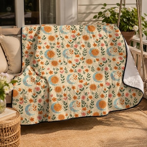 May include: A quilted throw blanket featuring a repeating pattern of smiling suns, crescent moons, and small pink and orange flowers on a cream background. The blanket has a white underside and a black stitched border along the edges.