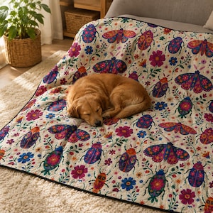Op de afbeelding: Een crèmekleurige, doorgestikte plaid met een herhalend patroon van kleurrijke kevers, motten en bloemen in roze, blauwe, oranje en paarse tinten. De deken is afgewerkt met een zwarte rand en heeft een zachte, textuurrijke stoffen afwerking.