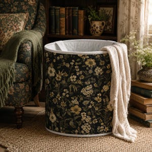 May include: A cylindrical laundry hamper featuring a dark black background with a dense, repeating pattern of cream, yellow, and muted green wildflowers. The interior is lined with white fabric, and a cream-colored knit throw blanket is draped over the side.