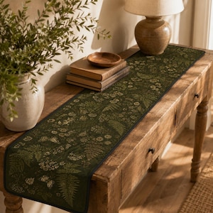 Può includere: Un runner da tavola lungo e rettangolare con uno sfondo verde scuro e una stampa botanica dettagliata di felci, piccoli fiori bianchi e farfalle. Il runner presenta un sottile bordo nero ed è disteso su una console in legno.