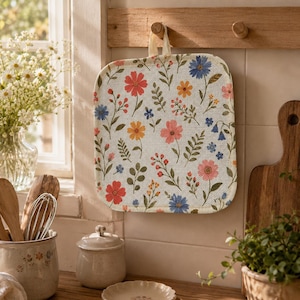 Könnte beinhalten: Ein quadratischer Topflappen aus Stoff mit einem wiederkehrenden Muster aus bunten Wildblumen, darunter rosa, orange, rote und blaue Blüten mit grünen Blättern und Stängeln auf cremefarbenem Grund, versehen mit einer Stoffschlaufe zum Aufhängen.