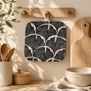 May include: A square fabric pot holder featuring a repeating black ginkgo leaf pattern on a cream-colored background. The pot holder has a small fabric loop at the top center for hanging and is suspended from a wooden wall-mounted hook rack.