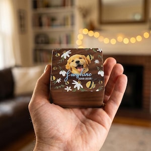 May include: A wooden box with a smiling golden retriever portrait, floral and bee accents, and the name "Sunshine" with the years 2008-2026. The box is held in a hand.
