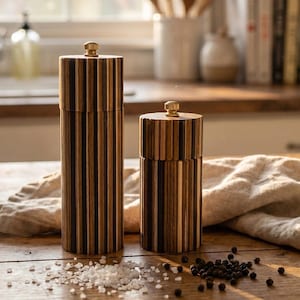May include: Two wooden salt and pepper grinders with a striped design. The grinders are made of alternating dark and light wood and have a gold-colored knob on top. Salt and peppercorns are scattered on the wooden surface.