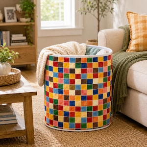 May include: A cylindrical storage basket featuring a vibrant, multi-colored patchwork grid pattern of small squares in red, blue, green, yellow, and brown. The basket has a white rim and contains a folded cream-colored textured fabric blanket.