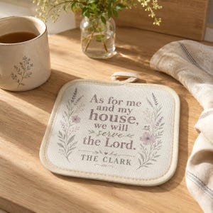 May include: A square fabric pot holder with a cream-colored background, featuring a floral border and the printed text: As for me and my house, we will serve the Lord. THE CLARK. It includes a small fabric loop at the top for hanging purposes.