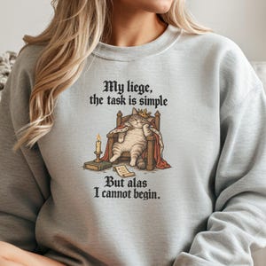 May include: A light gray crewneck sweatshirt featuring a graphic of a tabby cat wearing a crown and cape, sitting on a throne. The text reads: My liege, the task is simple. But alas I cannot begin. A candle on a book and a scroll are nearby.
