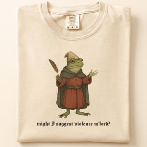 May include: Cream-colored t-shirt featuring a whimsical illustration of a frog dressed as a medieval figure, holding a quill. The frog wears a red robe, green cape, and a small hat. Text below reads: "might I suggest violence m'lord?"