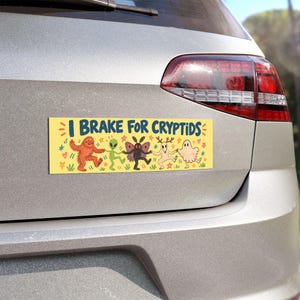 May include: A rectangular yellow bumper sticker with the blue text I BRAKE FOR CRYPTIDS. The design features a line of five illustrated creatures including a brown Bigfoot, a green alien, a dark moth-like figure, a deer-headed being, and a ghost.
