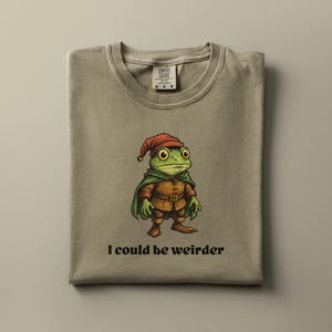 Puede incluir: Una camiseta Comfort Colors color beige con una rana de dibujos animados vestida de gnomo. La rana lleva un gorro rojo, una capa verde y un atuendo marrón. El texto "I could be weirder" está impreso debajo. La camiseta está doblada.