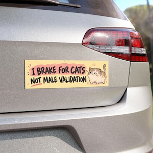 May include: A rectangular bumper sticker with a pale yellow background featuring a cartoon cat and the black text: I BRAKE FOR CATS NOT MALE VALIDATION. The text I BRAKE FOR CATS is highlighted by a pink brushstroke effect.