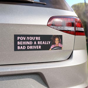 May include: A rectangular bumper sticker with a black background and light pink text that reads POV: YOU'RE BEHIND A REALLY BAD DRIVER. To the right of the text is a graphic of a doll with long brown hair wearing a pink top, holding a steering wheel.