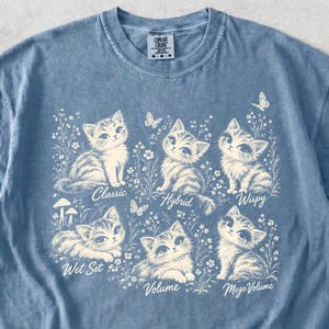 May include: A blue t-shirt featuring a cream-coloured graphic of five kittens surrounded by wildflowers and butterflies. The text labels Classic, Hybrid, Wispy, Wet Set, Volume, and Mega Volume are printed beneath each respective kitten illustration.