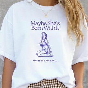 May include: A white short-sleeved t-shirt featuring a purple graphic print. The text reads "Maybe She's Born With It" above an illustration of a person kneeling, with the text "MAYBE IT'S ADDERALL" printed in purple below the illustration.