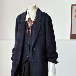 May include: A navy blue blazer, vest, and pleated skirt ensemble on a mannequin. The blazer has a two-button closure and the vest has a three-button closure. A white collared shirt with a patterned tie is visible. A brown backpack is held in the mannequin's hand.