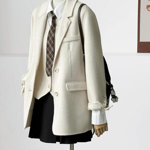 May include: Cream-colored blazer, vest, and skirt ensemble. The blazer has a classic two-button closure and notched lapels. A white collared shirt and a brown plaid tie are visible. A black skirt and a black backpack complete the outfit.