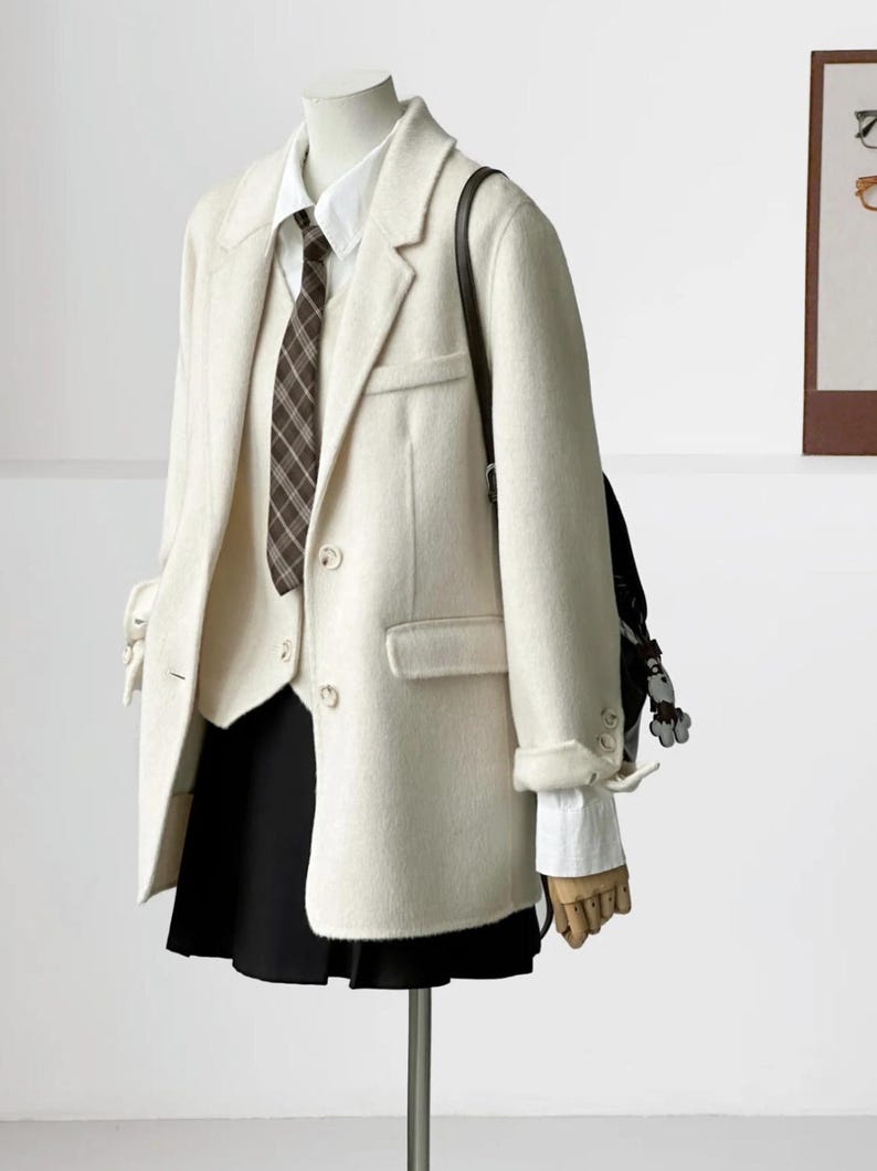 May include: Cream-colored blazer, vest, and skirt ensemble. The blazer has a classic two-button closure and notched lapels. A white collared shirt and a brown plaid tie are visible. A black skirt and a black backpack complete the outfit.