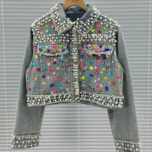 May include: A gray denim jacket adorned with faux pearls and colorful rhinestones. The jacket features a classic collar, two front pockets, and frayed edges. The cuffs and collar are embellished with pearls.