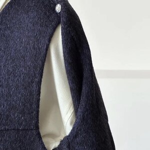 May include: Close-up of a navy blue wool coat with silver buttons. The coat has a unique design with an opening on the side, revealing a cream-colored lining. The fabric has a textured appearance.