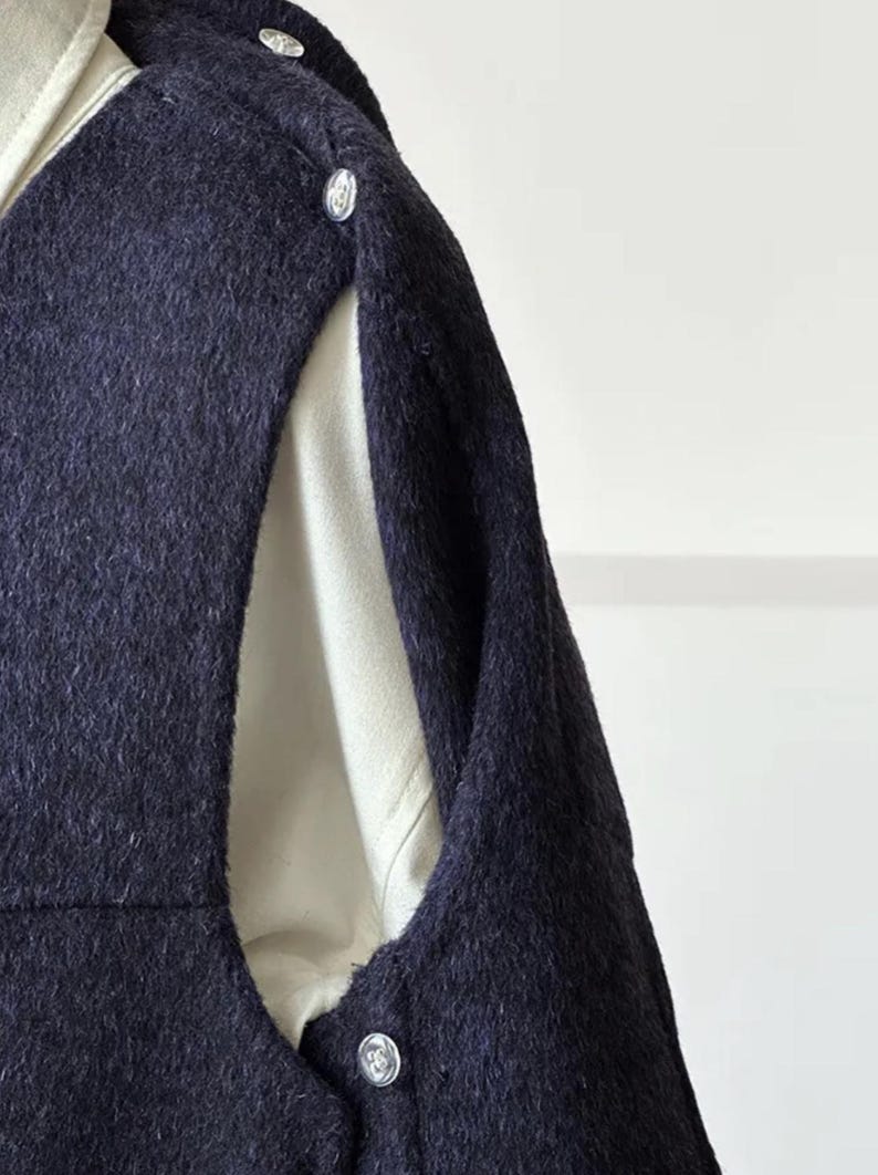 May include: Close-up of a navy blue wool coat with silver buttons. The coat has a unique design with an opening on the side, revealing a cream-colored lining. The fabric has a textured appearance.