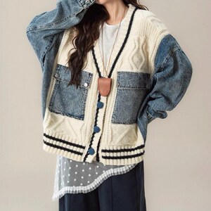 May include: A cream-colored cardigan with denim sleeves and pockets. The sweater features a cable knit design, black stripes at the bottom, and blue buttons. A brown leather pendant hangs from the neckline.