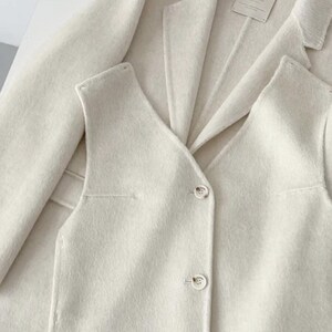 May include: Cream-colored blazer and vest set. The blazer has a classic lapel design, and the vest features a V-neck and button closure. The fabric appears soft and textured. The set is displayed on a white surface.